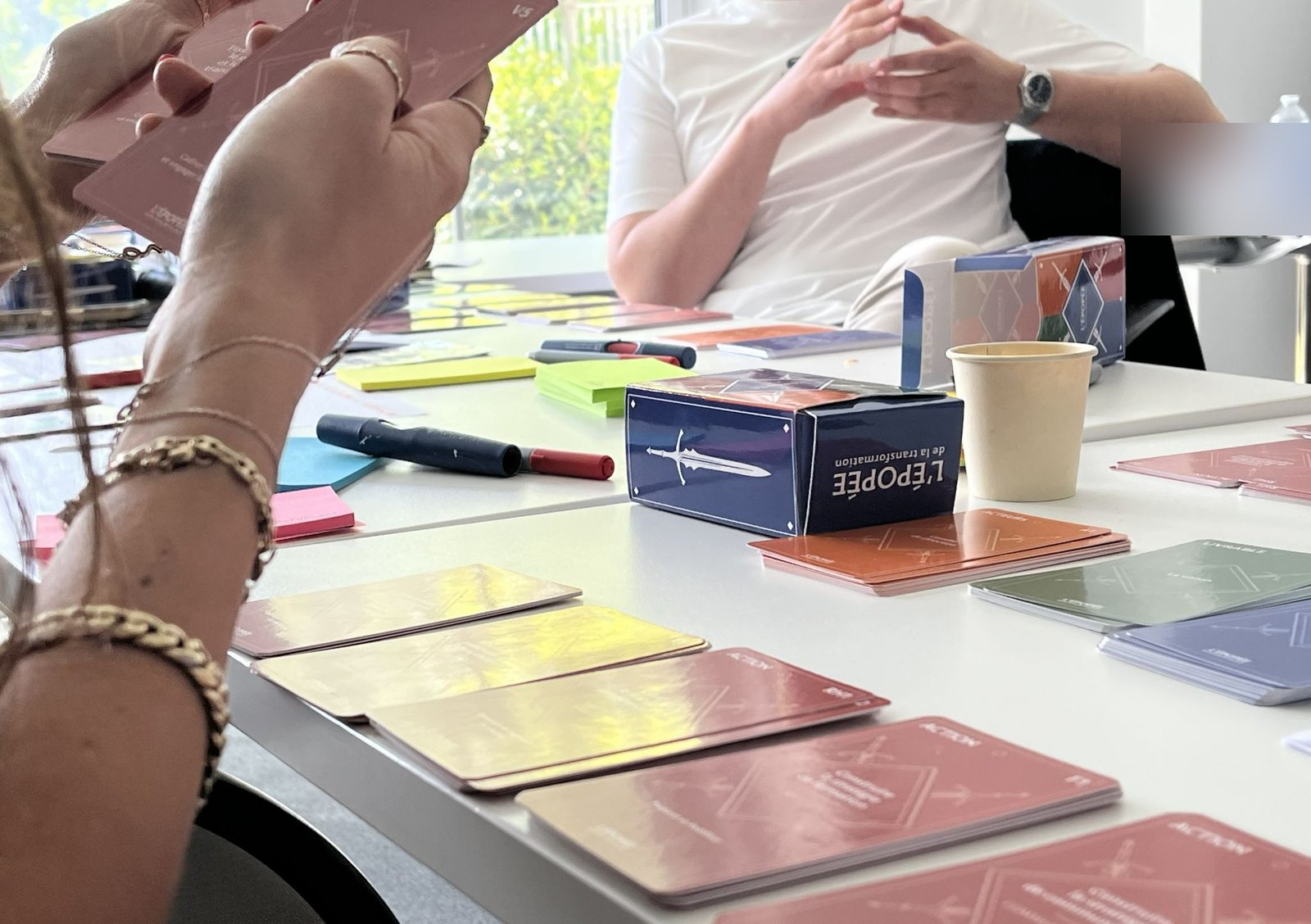 Des cartes ACTION de l'Épopée tenues en main au-dessus de la table, à côté de la boîte de jeu